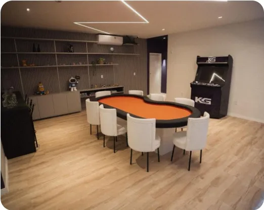 Sala com mesa de jogos laranja, cadeiras brancas, estante e fliperama com o logo KG
